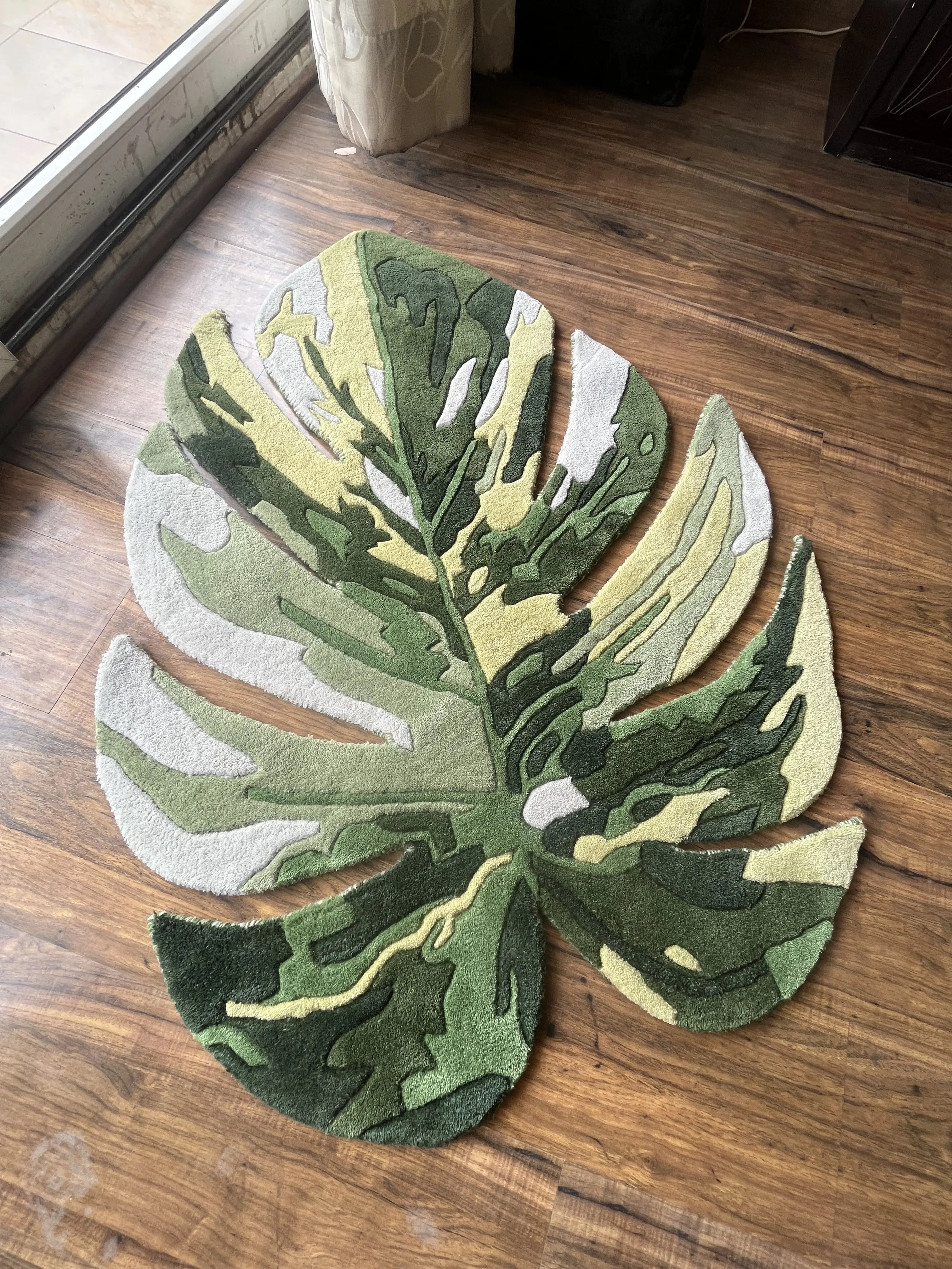 A rectangular area rug featuring a leaf pattern with green, yellow, and white shades, made of wool, placed on a wooden floor.
